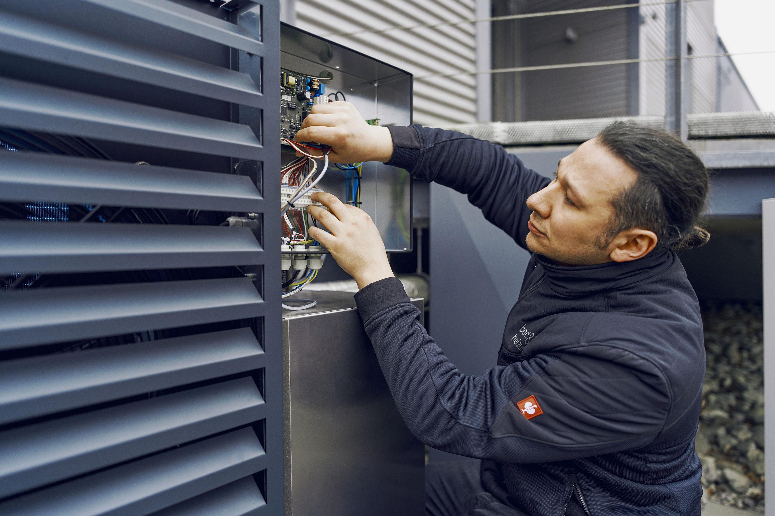 Ein Techniker überprüft eine Elektroinstallation an einer Außeneinheit, während er sorgfältig Kabel in einem offenen Schaltkasten anschließt.