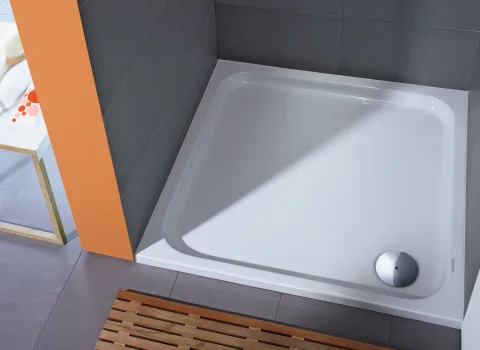 Eckige Duschwanne mit glatter, glänzender Oberfläche, eingefasst von grauen Fliesen und einer auffälligen orangefarbenen Wand.