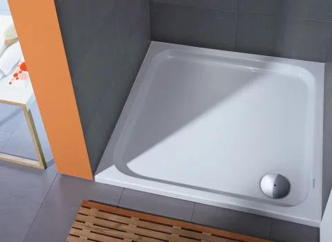 Eckige Duschwanne mit glatter, glänzender Oberfläche, eingefasst von grauen Fliesen und einer auffälligen orangefarbenen Wand.