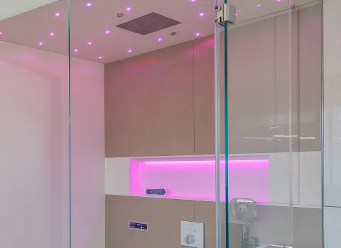 Das Bild zeigt eine moderne Dampfdusche mit Glaswänden, pinker LED-Beleuchtung an der Decke und einem beleuchteten Regal in der Rückwand.