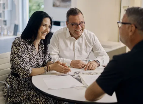 Zwei zufriedene Kunden unterhalten sich mit einem Badberater in einem modernen Büro. Vor ihnen liegen Unterlagen, während sie entspannt und lächelnd die Beratung genießen.
