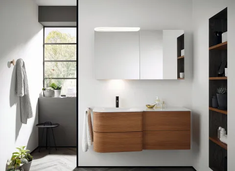 Ein helles Badezimmer mit einem wandmontierten Burgbad Badu-Waschtisch aus Holz und einem großen, beleuchteten Spiegel darüber.