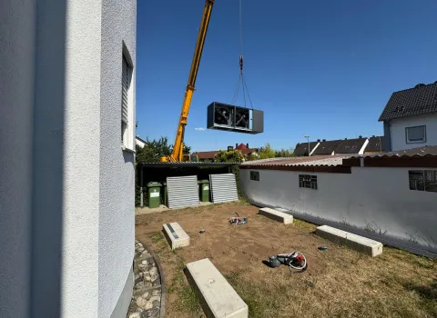 Kran hebt großes Außengerät einer Wärmepumpe über den Hof eines Mehrfamilienhauses.