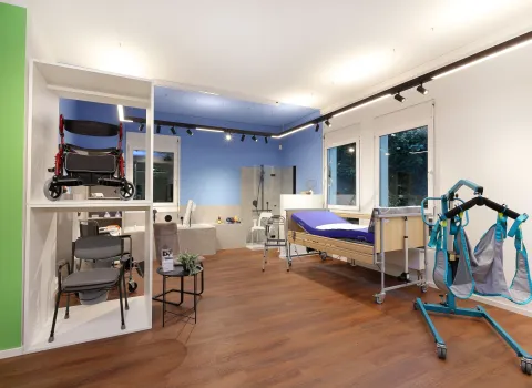 Blick in einen barrierefreien Schauraum mit farbigen Wänden in Grün und Blau, Pflegebett, Duschbereich und Reha-Hilfsmitteln.
