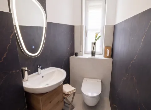 Modernisiertes Gäste-WC in Hersbruck mit Wand-WC, Waschtisch, Holz-Badmöbel und beleuchtetem Spiegel.