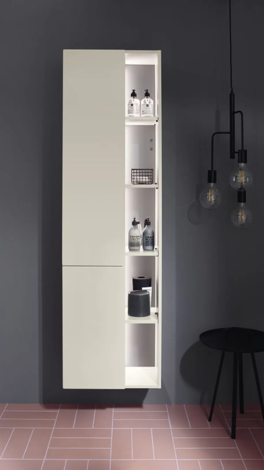 Der schlanke Burgbad Badu Hochschrank im Badezimmer ist halb geöffnet und zeigt beleuchtete Fächer, die Pflegeprodukte wie Seifenspender und Kerzen beinhalten.