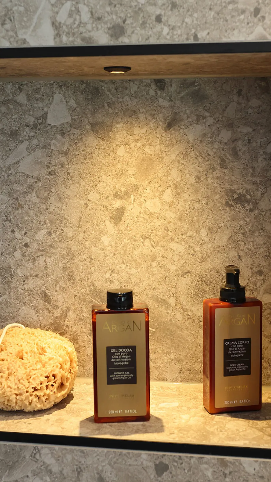 Eine beleuchtete Wandnische in einer Dusche aus Naturstein beherbergt zwei elegante Pflegeprodukte in bernsteinfarbenen Flaschen sowie einen natürlichen Badeschwamm.