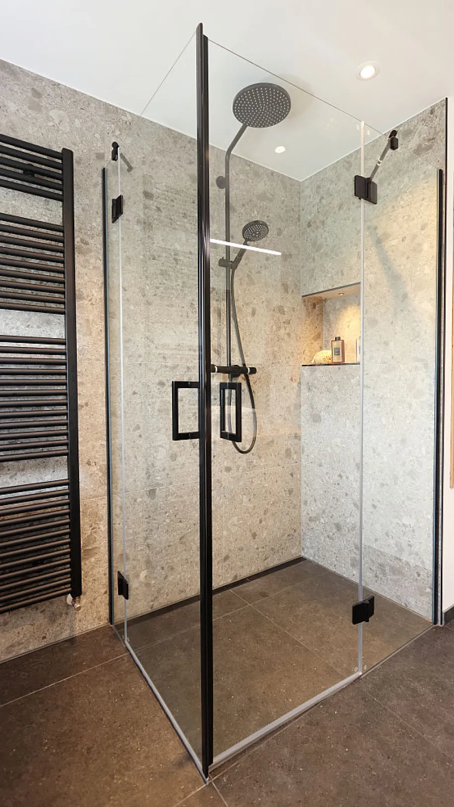 Eine begehbare Dusche mit Glastür und schwarzer Armatur ist in eine Wand aus grauen Steinfliesen eingelassen.