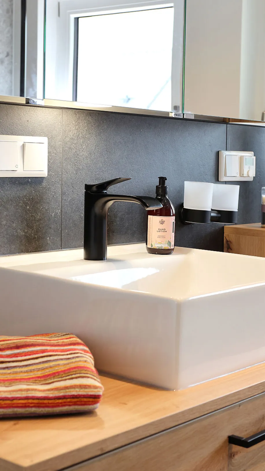 Ein stilvoll eingerichtetes Badezimmer zeigt ein eckiges, weißes Aufsatzwaschbecken mit schwarzer Armatur auf einer Holzplatte.